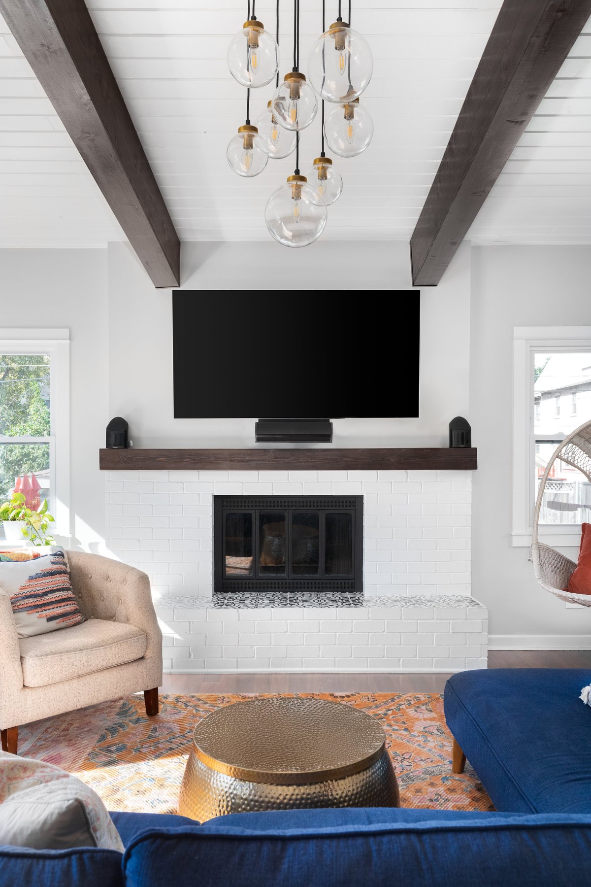 A renovated modern farmhouse living room with a blue couch, white fireplace, wood beams on a white shiplap ceiling, and a television mounted above the fireplace.