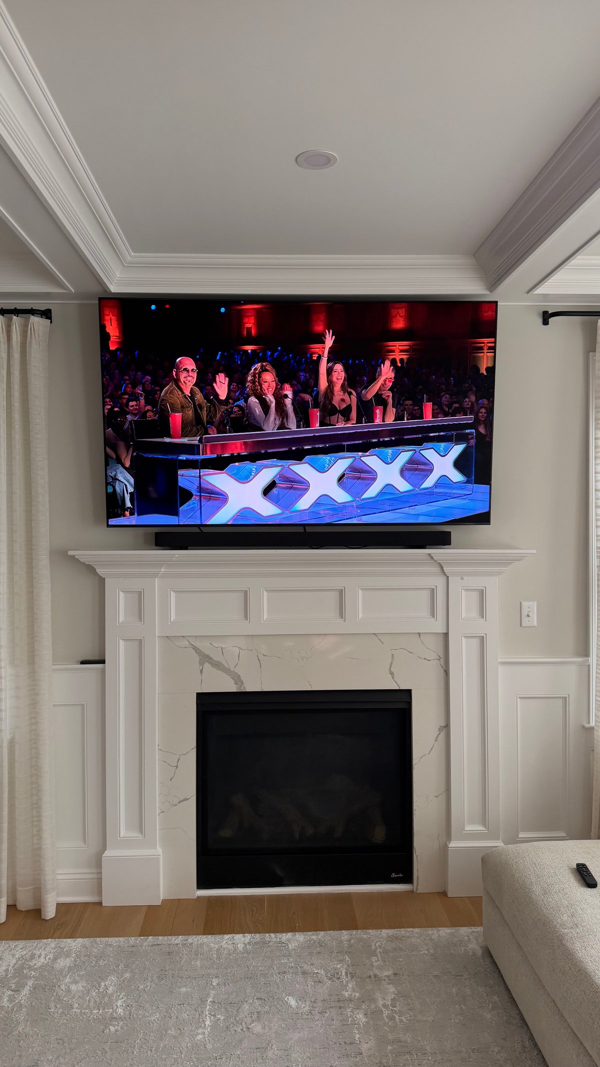 Flat-screen TV mounted above white fireplace mantel in living room, displaying talent show judges panel