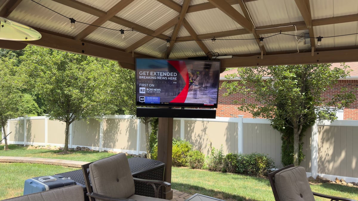 Outdoor patio with mounted TV under metal roof structure, surrounded by green lawn and white fence walls
