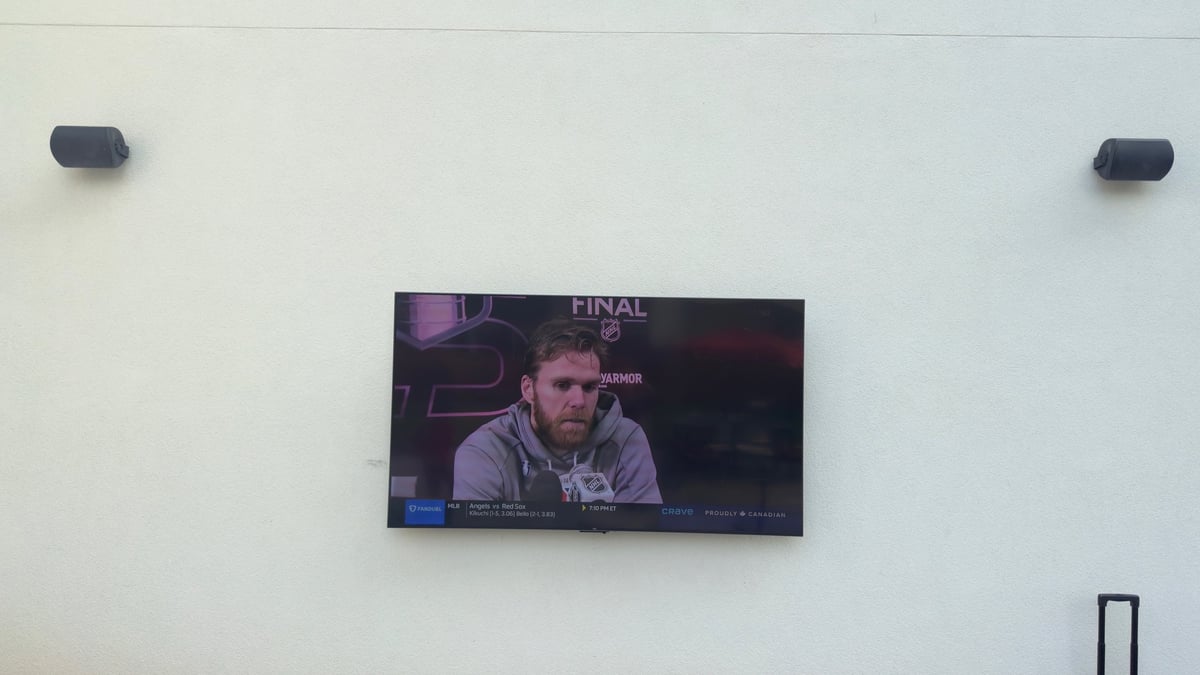 Wall-mounted TV displaying a mans face, with two speakers mounted above on white wall