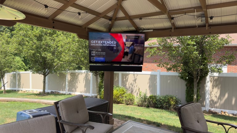 Outdoor covered patio area with chairs and TV screen mounted on wooden structure, green lawn and trees visible in background