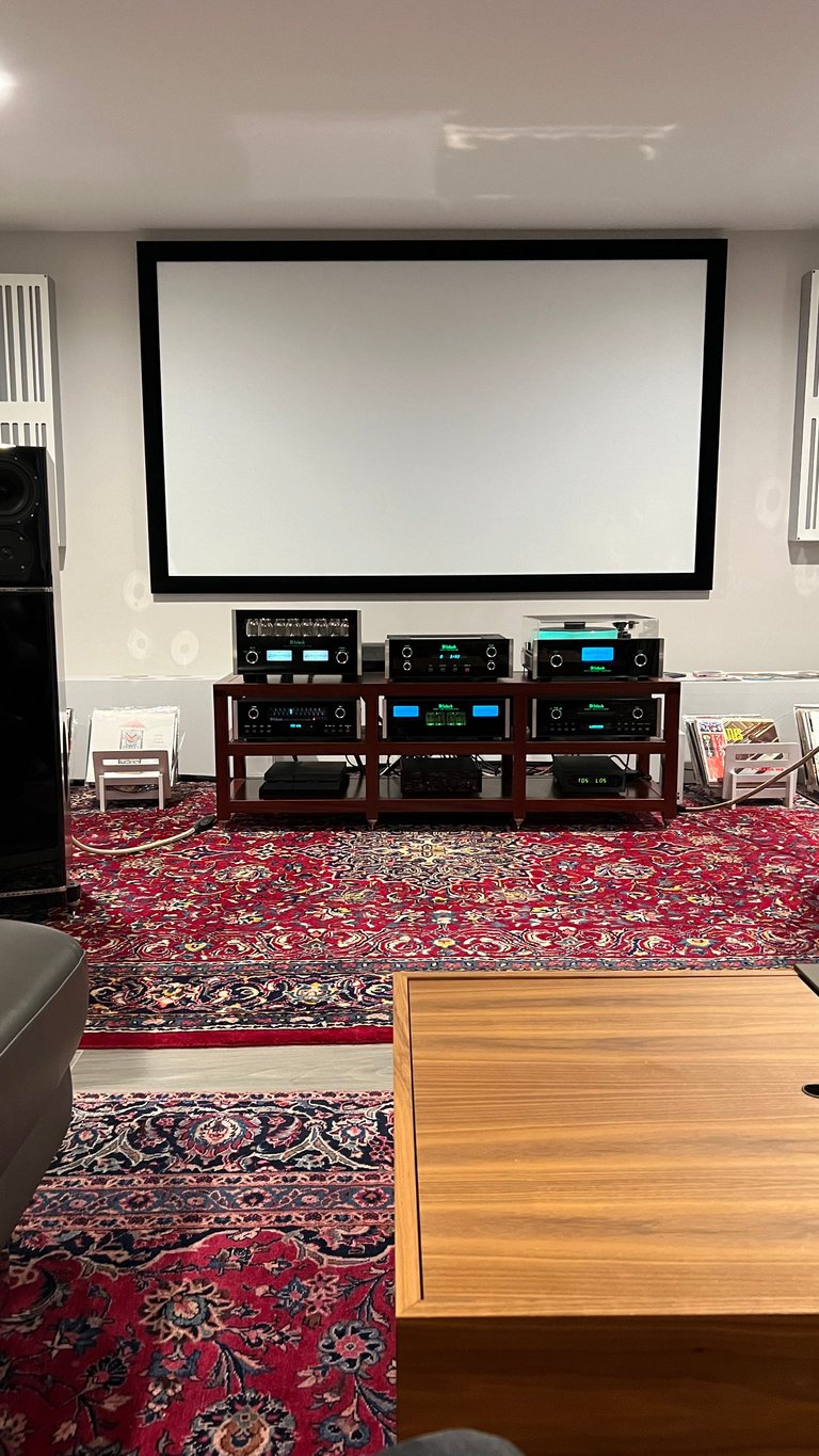 Home theater room with large projection screen, audio/video equipment on shelves, ornate red rug, and wooden furniture