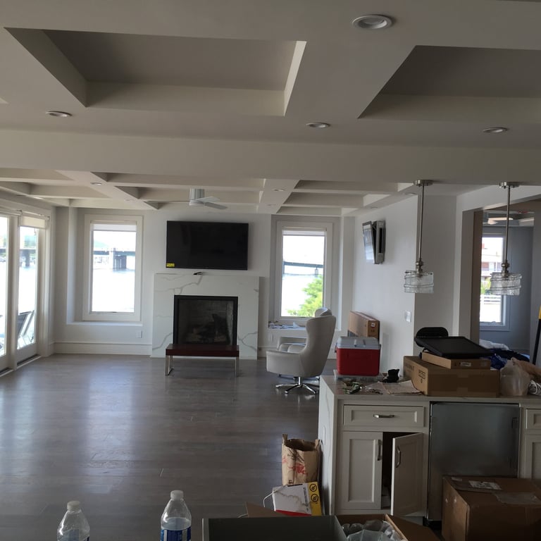 Spacious open-concept living room with fireplace, mounted TV, coffered ceiling, multiple windows, and kitchen area with boxes during move-in preparation