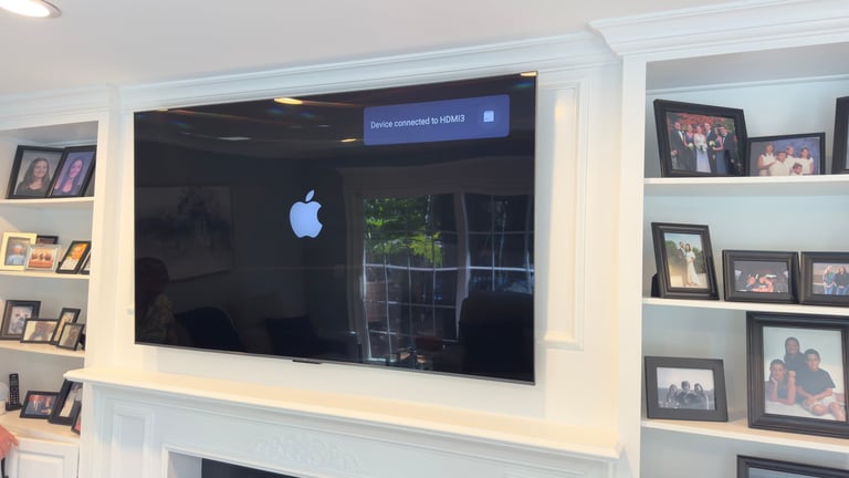 Large flat-screen TV mounted on white wall displaying Apple logo, flanked by shelves with framed photos